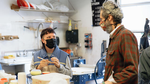 Instructor addresses student sitting at table with hand made ceramic item holding a paintbrush