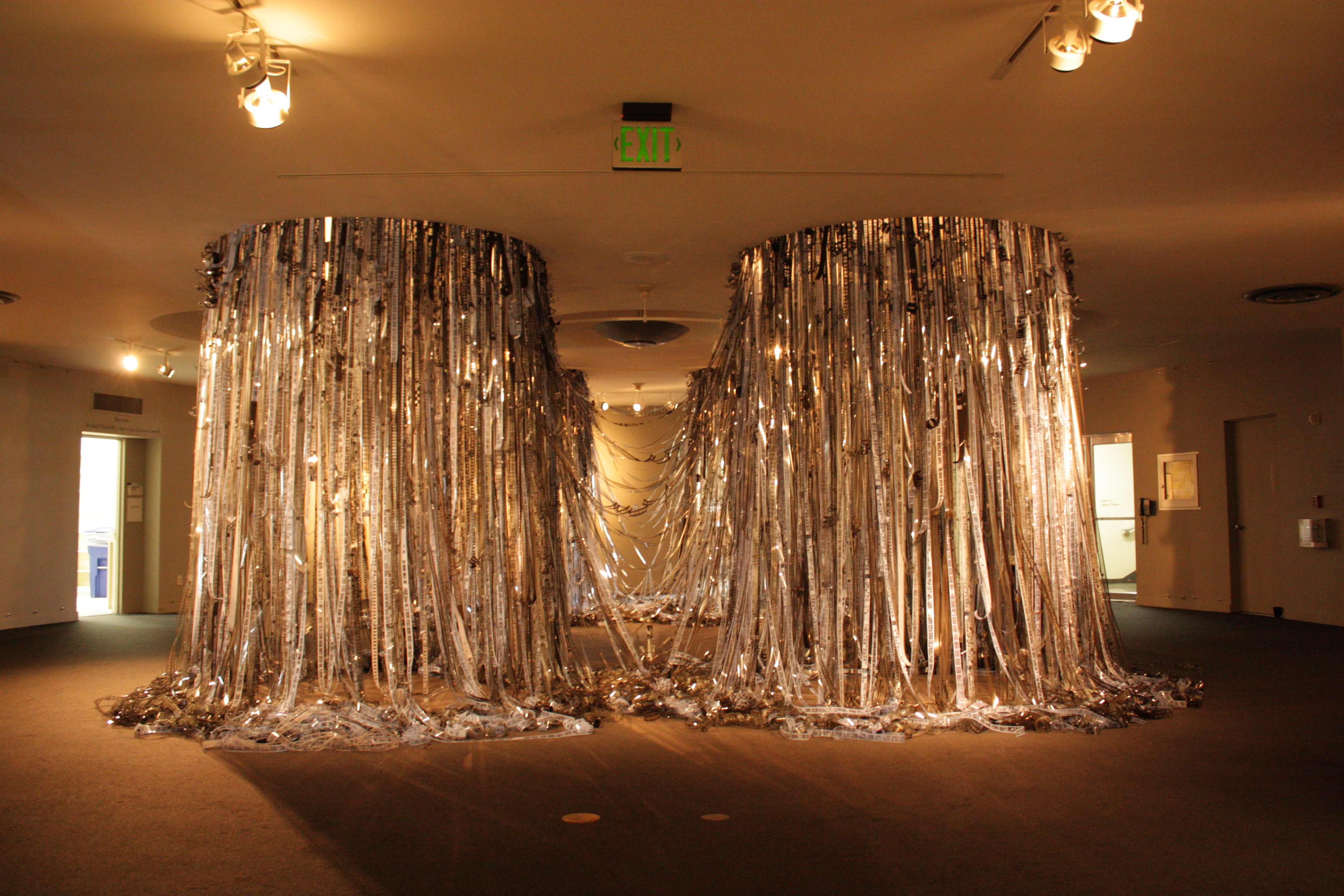 An art installation featuring cascading strands of reflective materials hanging from the ceiling, forming two large, curtain-like structures with light reflecting off their surfaces.