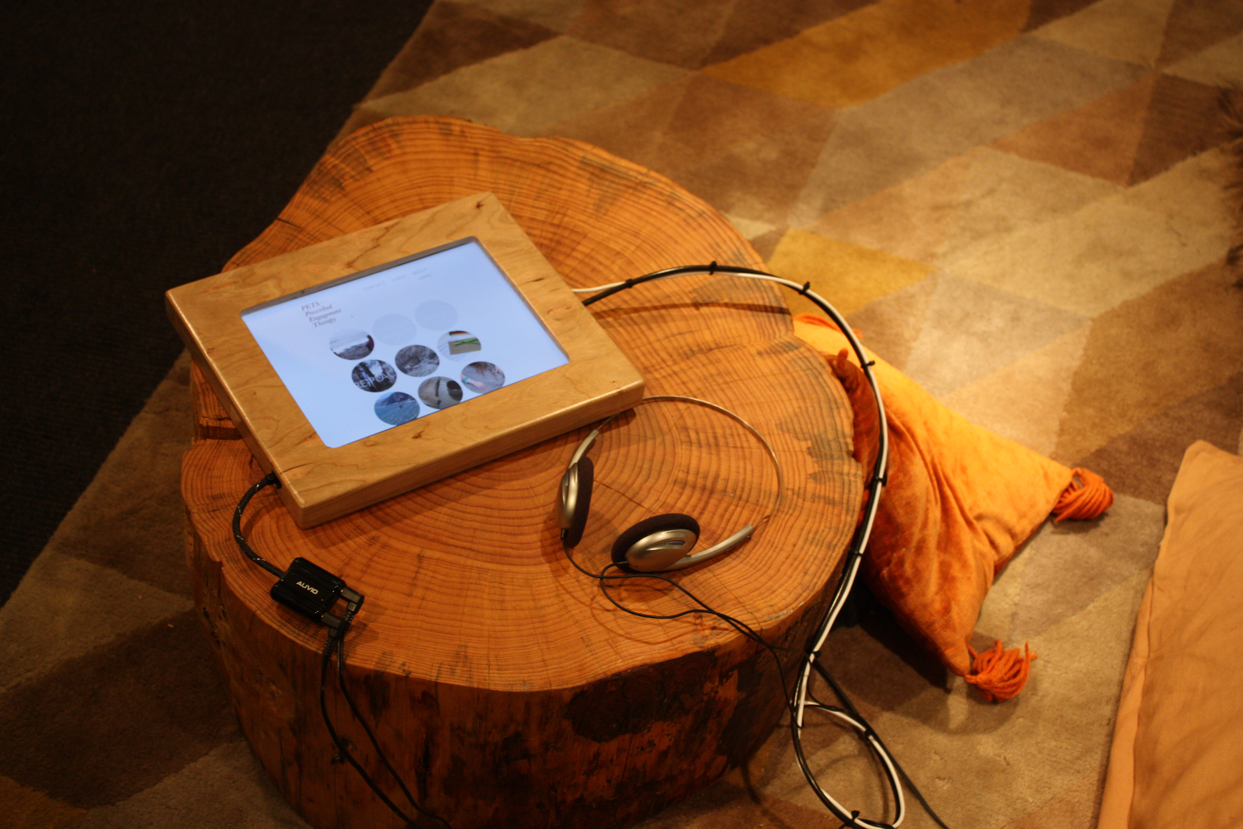 A digital tablet encased in a wooden frame is placed on a polished tree stump, accompanied by headphones, creating a cozy multimedia setup with pillows in the background.