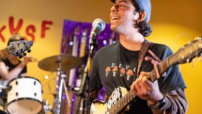 Student playing a guitar sings into a microphone in front of a hand made sign that reads KUSF