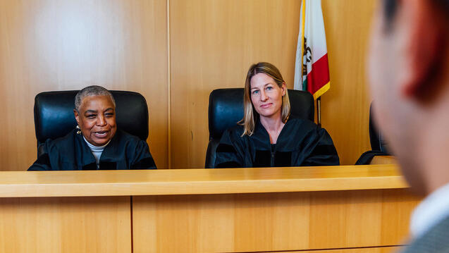Two judges behind their desk speak to a student in moot court.