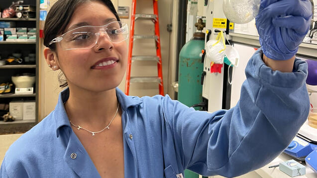 Student in chemistry lab holds up a beaker and looks at contents.