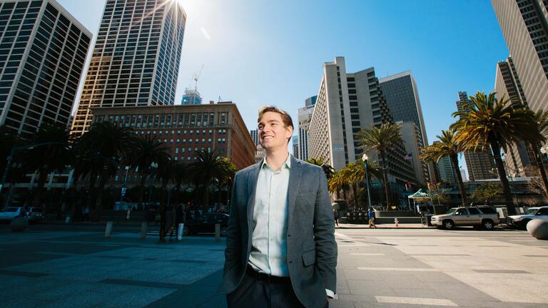 Student walks down the street with SF skyline behind him.