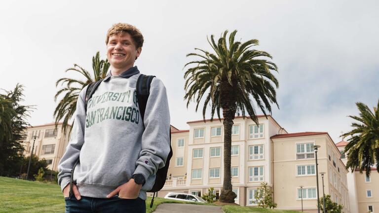 Student in USF sweatshirt stands in front of the Lone Mountain dorms.