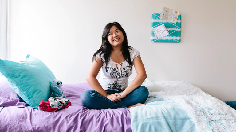 Student sitting on bed in residence hall, smiling