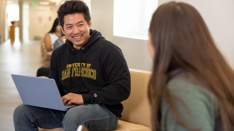 A student wearing a University of San Francisco hoodie smiling and in conversation with another person