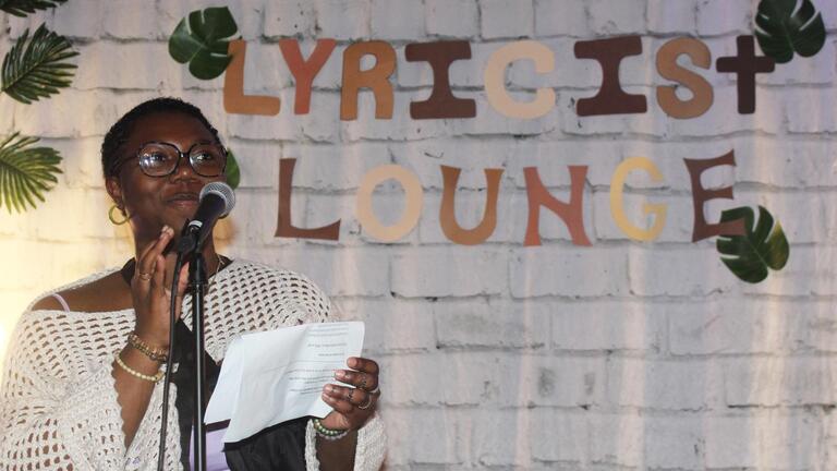 Lyricist Lounge host at the mic in front of a white brick background