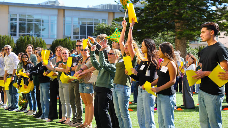 students, alumni, faculty, and staff hold up yellow papers to help form the shape of the letters CAS