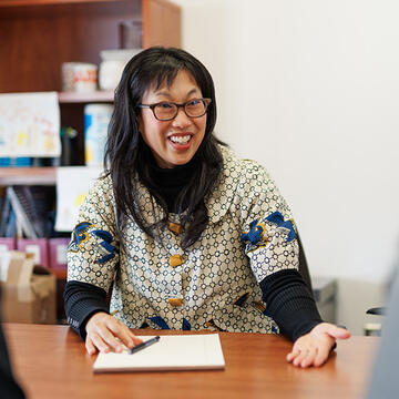 Person with glasses and patterned blouse smiling and gesturing to people sitting across the table